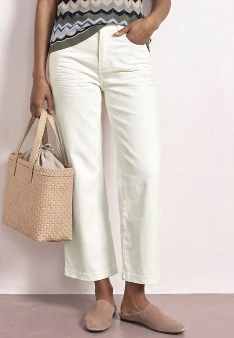 Eine Frau trägt cremefarbene, weite Cropped-Hosen, flache Pantoletten und hält eine gewebte Tasche.