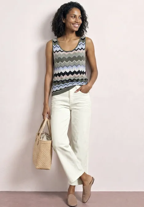 Eine Frau mit schwarzen lockigen Haaren trägt einen gemusterten Tanktop, helle Hosen, Basttasche und flache Schuhe.