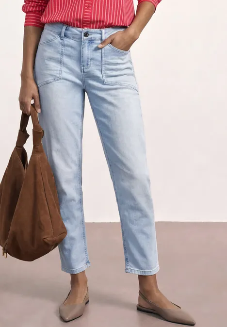 Eine Person in hellblauen Jeans, roter Bluse, hellbraunen Slippern und brauner Tasche vor hellem Hintergrund.