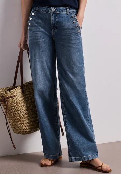 Une femme porte un jean large, des sandales et tient un sac en paille tressée.