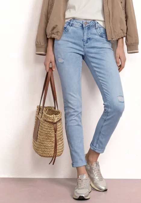 Person in hellblauen Jeans, beiger Jacke und Sneakern hält eine geflochtene Strohtasche.