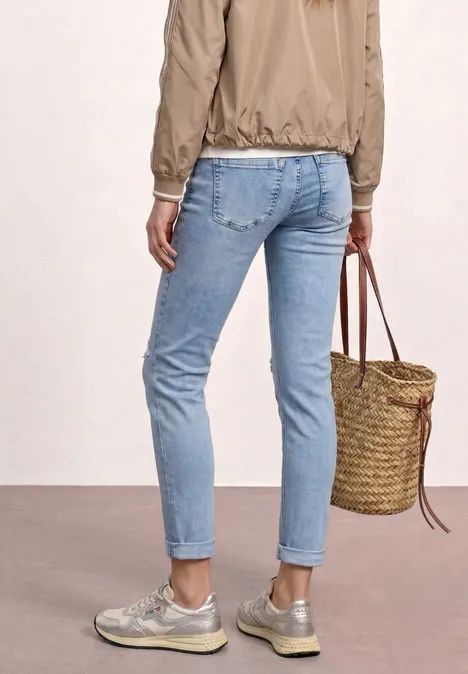 Rückenansicht einer Person in hellblauen Jeans, silbernen Sneakers, beiger Jacke und geflochtene Korbtasche.