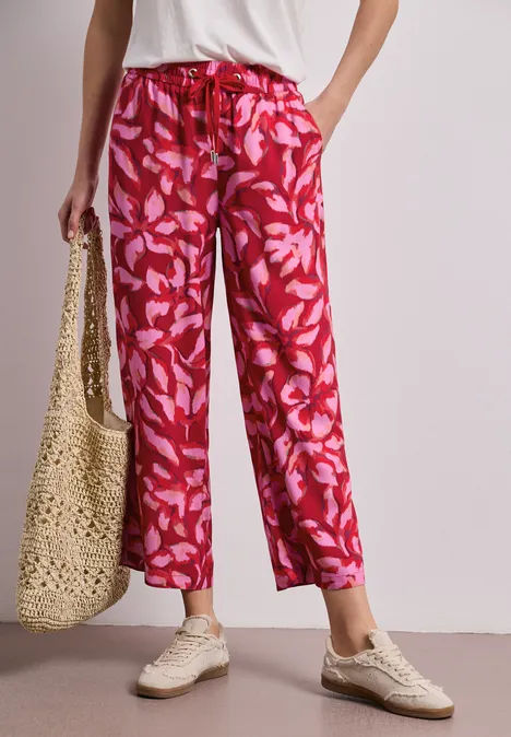 Vrouw draagt rood, wijd vallende bloemensbroek, witte sneakers en een strooien tas.