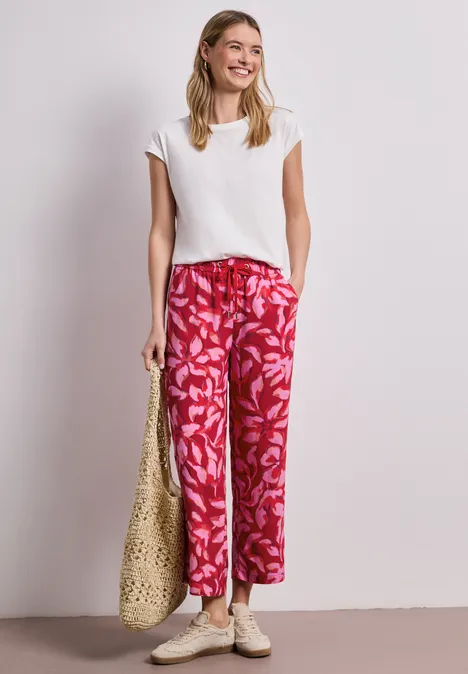 Vrouw in witte top en rode, gebloemde wijde broek, met rieten tas en sandalen tegen neutrale achtergrond.