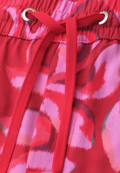 Rode stof met roze bloemenprint en rode koordjes door zilverkleurige ringen.