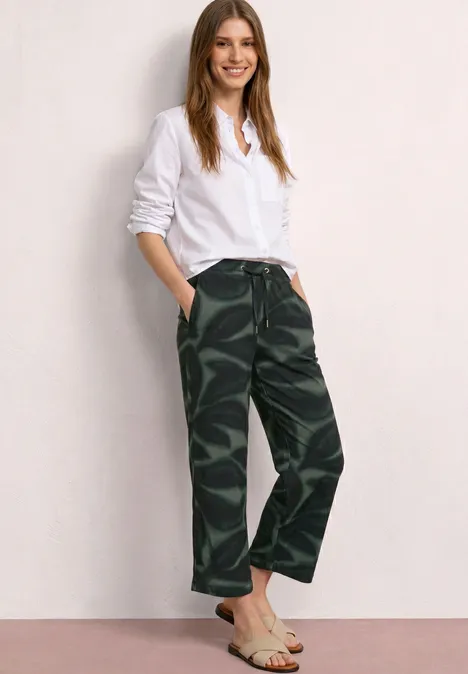 Eine Frau in weißem Hemd und Camouflage-Hose lehnt vor einer hellen Wand.