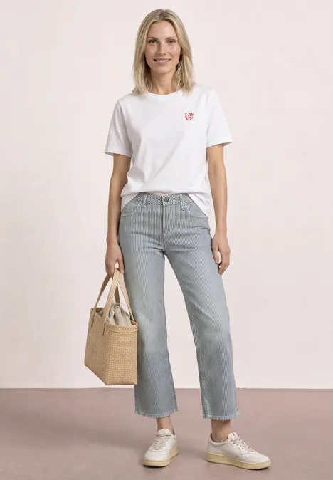 Eine Frau mit weißem T-Shirt, heller Jeans, Sneakers und Korbtasche steht vor hellem Hintergrund.