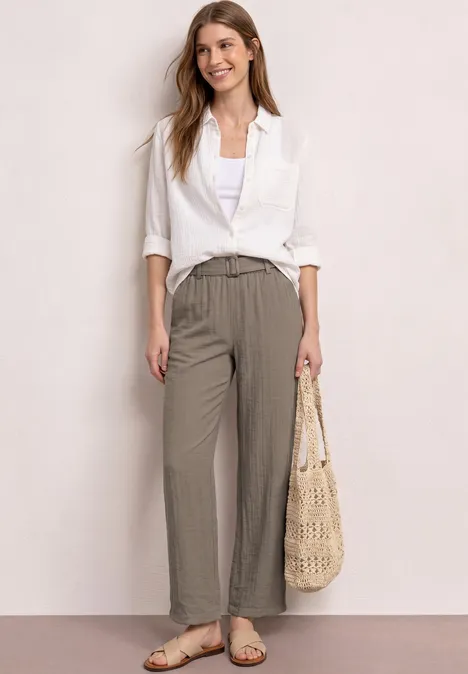 Frau in weißer Bluse, weiten taupefarbenen Hosen und Sandalen, hält einen geflochtenen Korbbeutel.