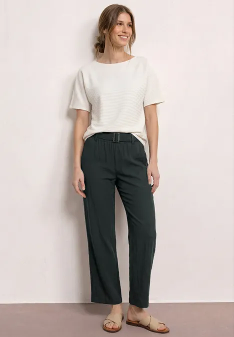 Frau in weißem T-Shirt, dunklen Hose mit Gürtel und Sandalen vor hellem Hintergrund.