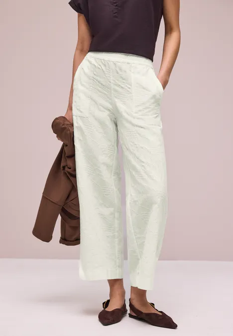 Vrouw in lichte, wijde broek, donker topje ingestopt, met een bruin jasje om de arm en bruine loafers.