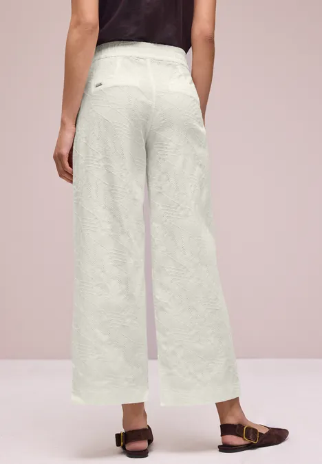 Rugaanzicht van een persoon in wijde lichtgekleurde linnen broek en bruine sandalen tegen neutrale achtergrond.