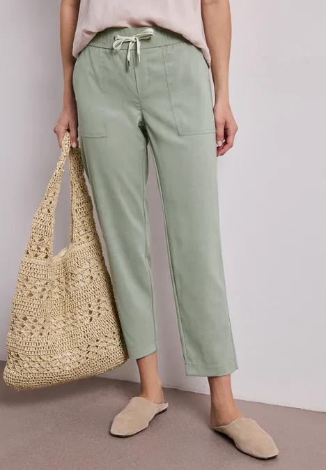 Persoon draagt lichtgroene, rechte broek, beige top, rieten tas en comfortabele instappers.