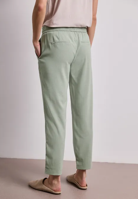 Persoon van achteren, draagt lichtgroene joggingbroek en slippers, handen in de zakken.