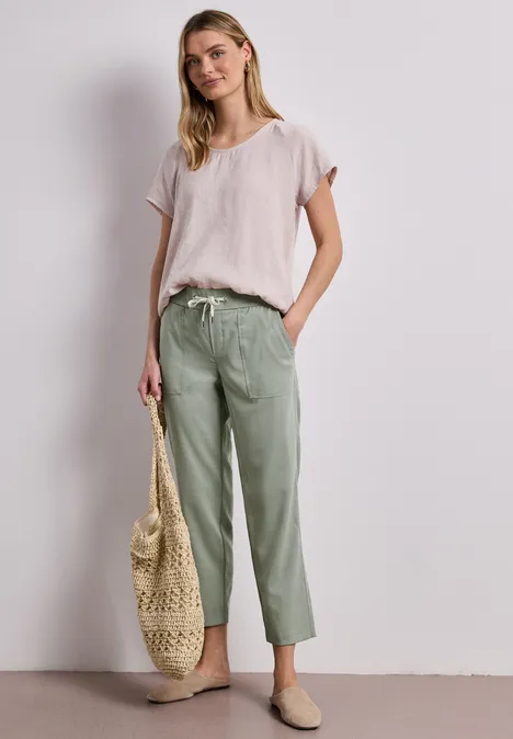 Vrouw in los lichtroze T-shirt en groene linnen broek, met strooien tas en instappers voor een neutrale achtergrond