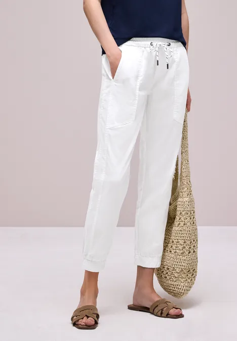 Witte linnen broek met koord en zakken, gecombineerd met sandalen en gevlochten rieten tas.