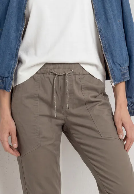 Eine Person trägt graubraune Kordelzug-Hose mit Eingrifftaschen, weißes T-Shirt und Jeansjacke.