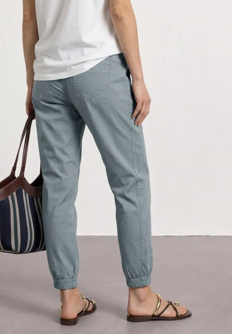 Slim legs broek in lichtgewicht kwaliteit washed blue Persoon van achteren in grijze broek met elastische taille, wit shirt, gestreepte tas en platte sandalen.