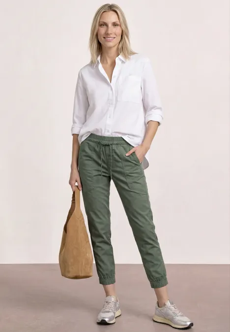 Eine Person trägt weiße Bluse, olivgrüne Hose, graue Sneaker und hält einen beigen Beutel.