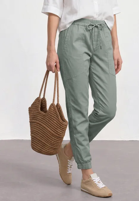 Slim legs broek in een lichtgewicht stof dark mint green Vrouw in groene joggerbroek met elastische boorden, wit shirt, gevlochten tas en beige sneakers.