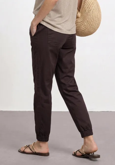 Slim legs broek in lichtgewicht kwaliteit ruby vine Donkerbruine, casual joggerbroek met elastische boorden, gecombineerd met sandalen, beige shirt en een strooien tas.