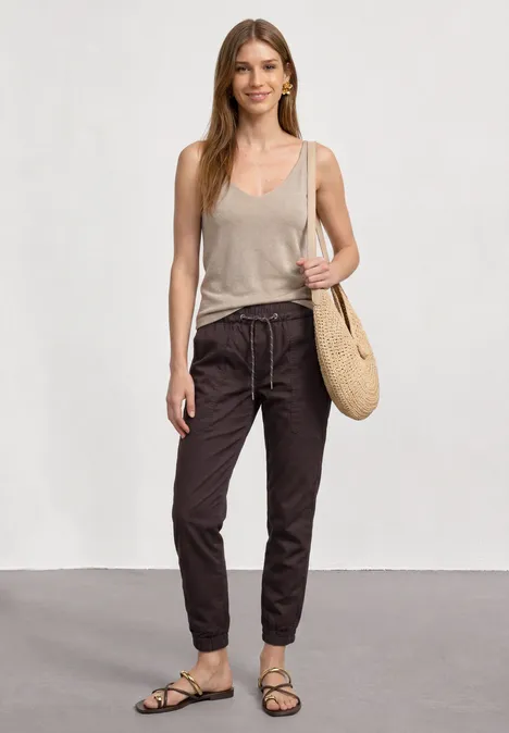 Slim legs broek in lichtgewicht kwaliteit ruby vine Vrouw draagt beige top, donkere broek en sandalen, met een strooien tas.