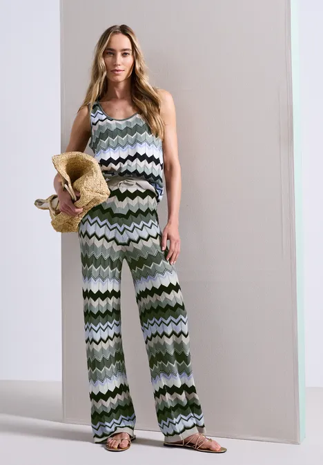 Vrouw in een mouwloze, groen-witte zigzag jumpsuit met wijde pijpen, houdend een strohoed vast.