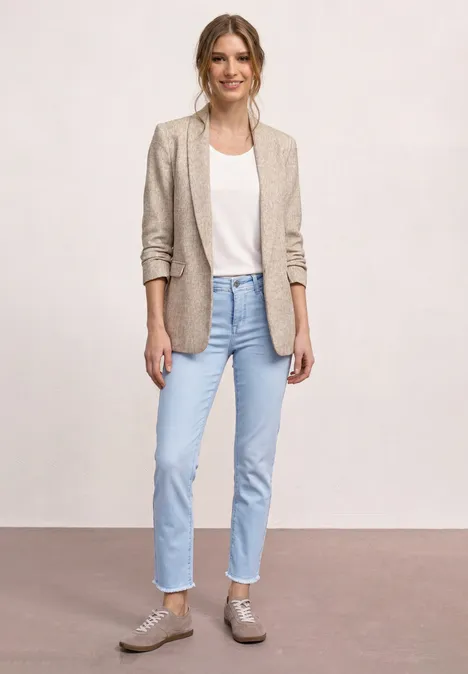 Eine Frau trägt beige Blazer, weißes T-Shirt, helle Jeans und graue Sneaker vor hellem Hintergrund.