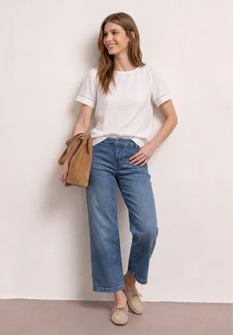 Vrouw in wit T-shirt, wijde cropped jeans en espadrilles, met beige schoudertas voor een lichte achtergrond.