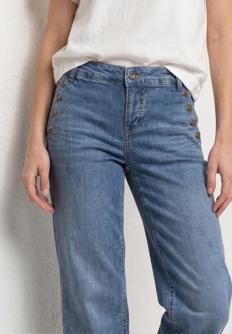 Detailfoto van iemand met een lichtblauwe, hoog getailleerde spijkerbroek en een wit shirt voor een lichte muur.