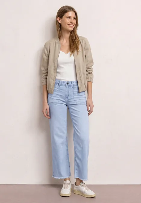 Vrouw draagt wit topje, beige bomberjack, wijde lichtblauwe jeans en lichte sneakers.