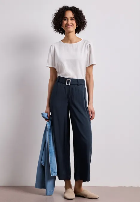 Frau im weißen T-Shirt und schwarzer High-Waist-Hose mit Gürtel, hält blaue Jacke.