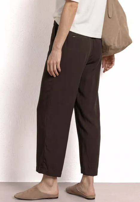 Zijaanzicht van persoon in een losse donkerbruine broek, beige instappers en een bijpassende tas.