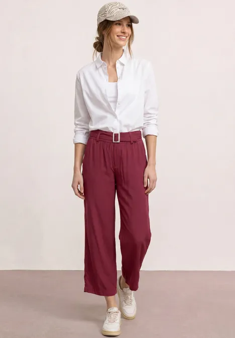 Eine Frau trägt weiße Bluse, weinrote Hose mit Gürtel und weiße Sneaker vor hellem Hintergrund.
