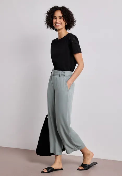 Frau mit lockigen Haaren trägt schwarzen T-Shirt, hellgraue Culottehose und schwarze Sandalen.