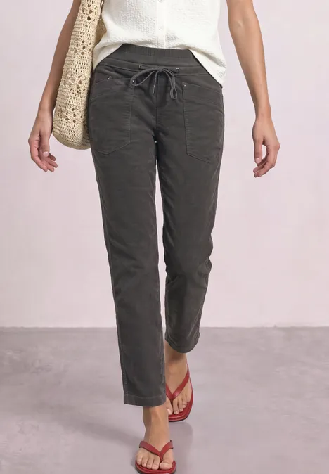 Person steht in dunkler, lässiger Hose, weißem Top, roten Flipflops und trägt eine Basttasche.