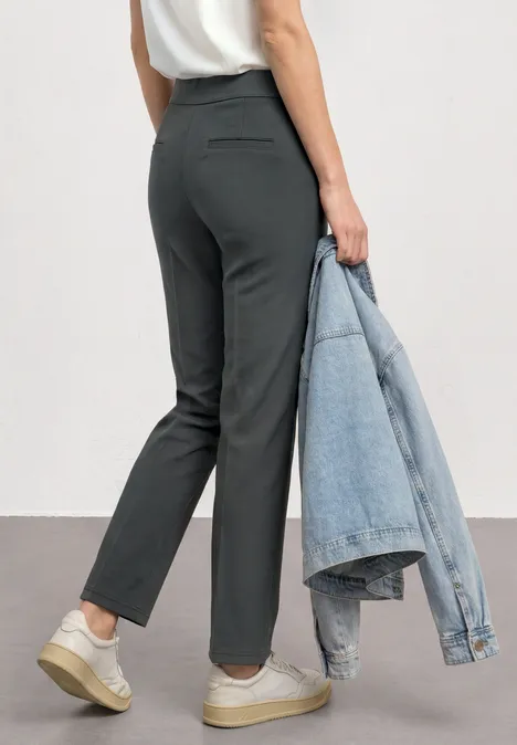 High Waist Straight Leg Hose im Casual Fit mossbound green Frau von hinten in dunkelgrauen Hosen und weißen Sneakern, die eine Jeansjacke in der Hand hält.