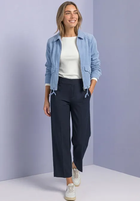 Femme en veste bleu clair, top blanc, pantalon large bleu marine et baskets sur fond neutre.
