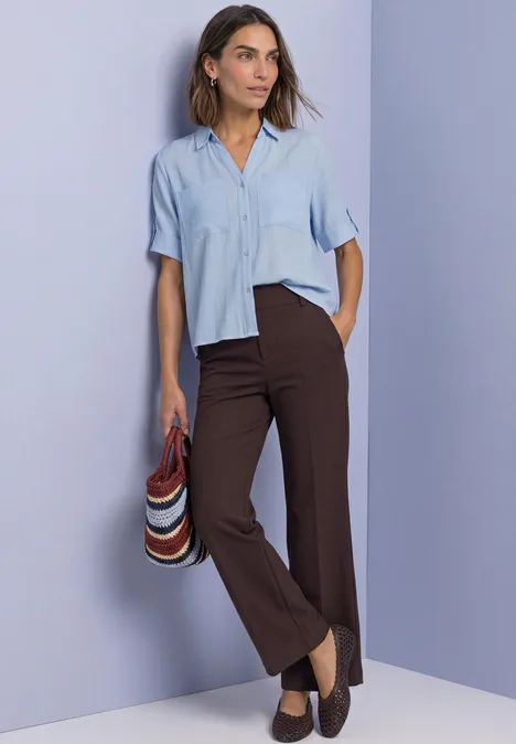 Femme en chemise bleue et pantalon marron porte un chapeau rayé devant un mur lilas