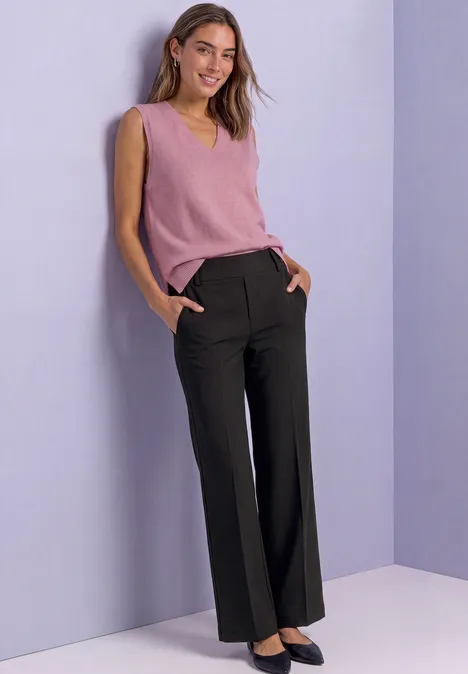 Eine lächelnde Frau in rosa Top und schwarzen Hosen lehnt an einer hellvioletten Wand.