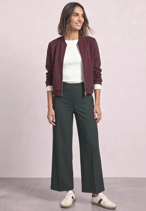 Femme portant un t-shirt blanc, un bomber bordeaux, un pantalon large gris foncé et des baskets blanches devant un fond clair