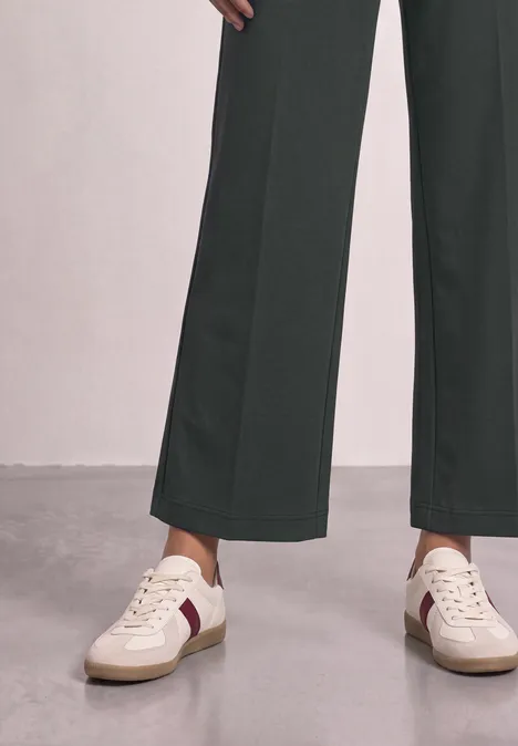 Deux jambes en pantalon ample foncé et baskets blanches à détails rouges posées sur un sol gris.