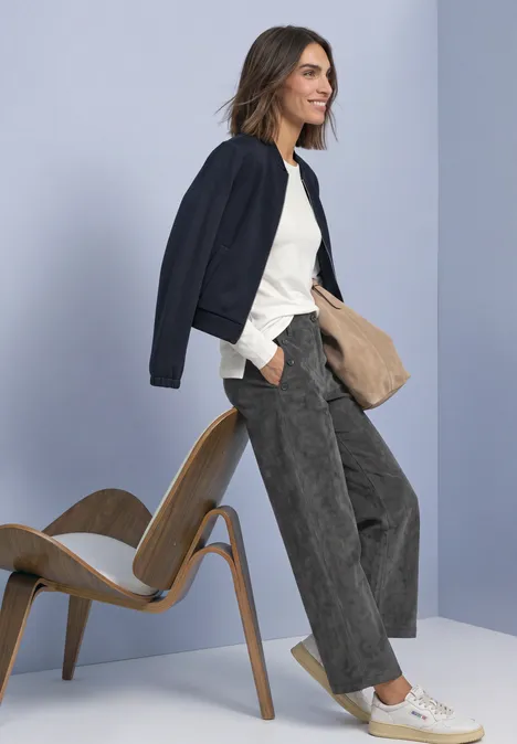 Frau im Casual-Look mit dunkler Jacke, weißem T-Shirt und weiten Cordhosen, neben Holzstuhl.