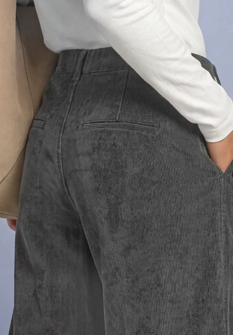 Eine Person in hoch taillierten schwarzen Jeans mit Hand in Tasche, weißem Oberteil und beigem Schulterriemen.