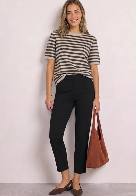 Frau in gestreiftem T-Shirt, schwarzen Hosen und brauner Tasche vor hellem Hintergrund