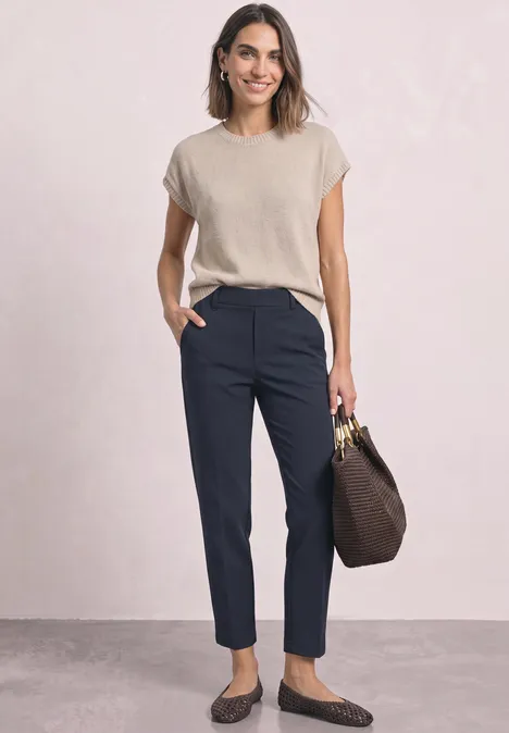 Frau in beigefarbenem Top, dunkelblauen Hosen und Mokassins, hält eine braune Handtasche.