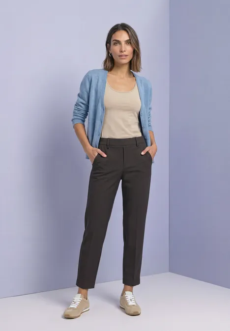 Eine Frau trägt helle Cardigan, beige Top und schwarze Hose vor lila Hintergrund.