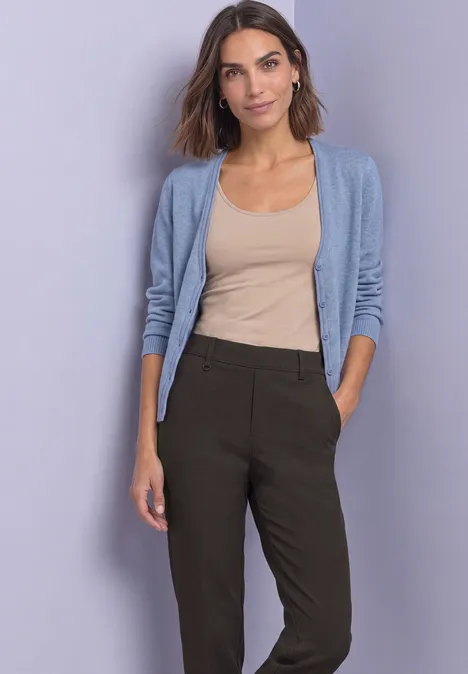 Frau in beigem Top, hellblauer Strickjacke und dunkler Hose vor schlichtem Hintergrund.