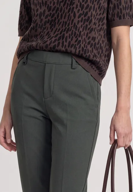 Frau in grünem Slim-Fit-Hose und braunem, gemustertem Oberteil, hält braune Tasche in der Hand.
