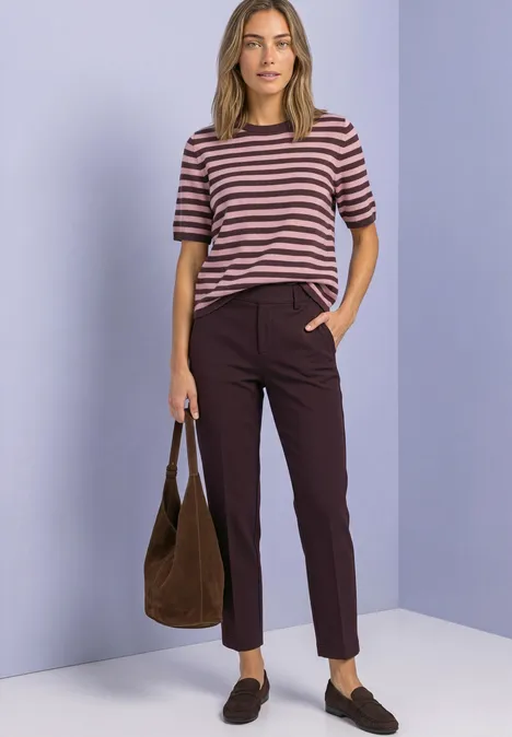 Eine Frau in gestreiftem T-Shirt, dunklen Hosen, braunen Loafers und Tasche steht vor lila Wand.
