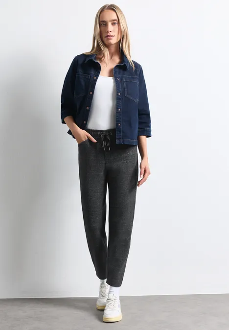 Frau steht vor weißer Wand, trägt Jeansjacke, weißes Top, graue Hose und weiße Sneaker.